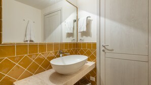 Une salle de bain avec un lavabo blanc, un pare-feu jaune et un miroir qui reflète une porte et des serviettes.