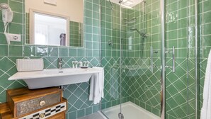 A bathroom with a white sink, green tiled walls, a glass shower enclosure, and a wooden chest of drawers.