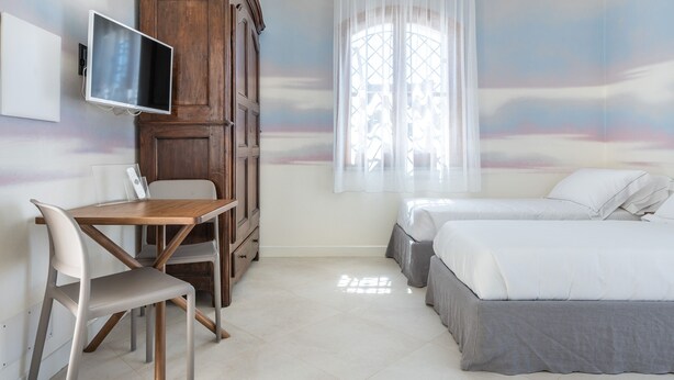 A bedroom with a wooden desk, two beds, a TV, and a window with sheer curtains.