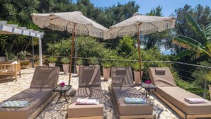 A sunlit outdoor lounge area with cushioned chairs and tables, surrounded by potted plants and a pergola with a fringe canopy. - PRESTIGE VILLAS 'Tree Tops' - North East Coast Villa with infinity pool... (Agios Markos, Corfu)