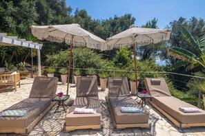 A sunlit outdoor lounge area with cushioned chairs and tables, surrounded by potted plants and a pergola with a fringe canopy. - PRESTIGE VILLAS 'Tree Tops' - North East Coast Villa with infinity pool... (Agios Markos, Corfu)