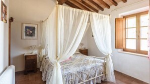 Een slaapkamer met een hemelbed, houten balken in het plafond, een raam met houten luiken en een lijst met een afbeelding aan de muur.