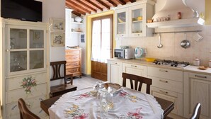 A kitchen with wooden beams, a dining table with a floral tablecloth, and a TV mounted on the wall. - Ancient farm la Martinella Depandance apartment (Siena)