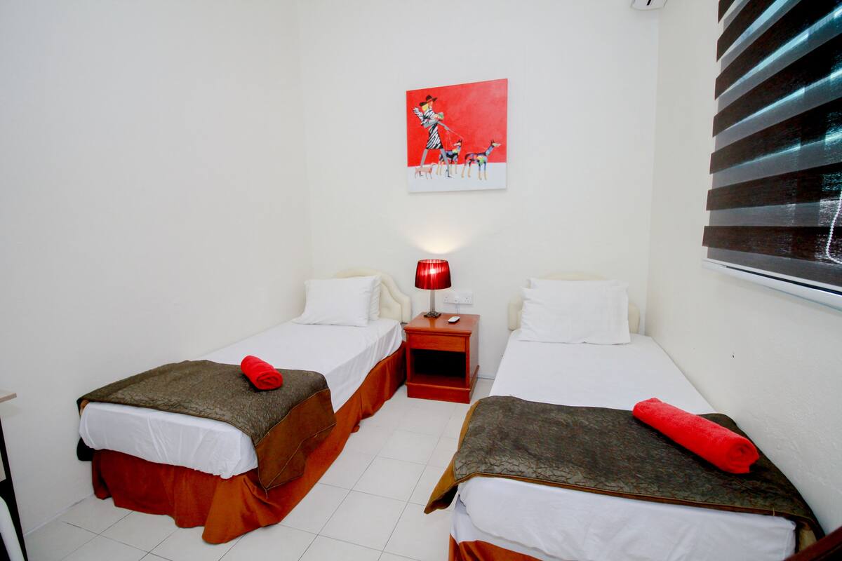 Habitación de hotel con dos camas individuales, una lámpara roja, un cuadro en la pared y una ventana con persianas.