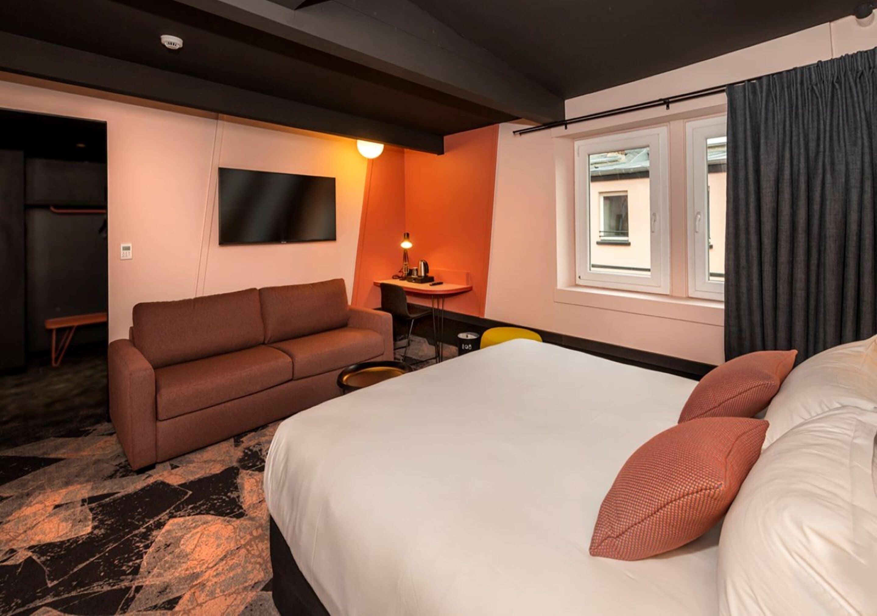 Una habitación de hotel con cama, sofá, televisión, escritorio y ventana con cortinas.