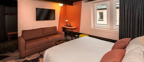 Una habitación de hotel con cama, sofá, televisión, escritorio y ventana con cortinas.