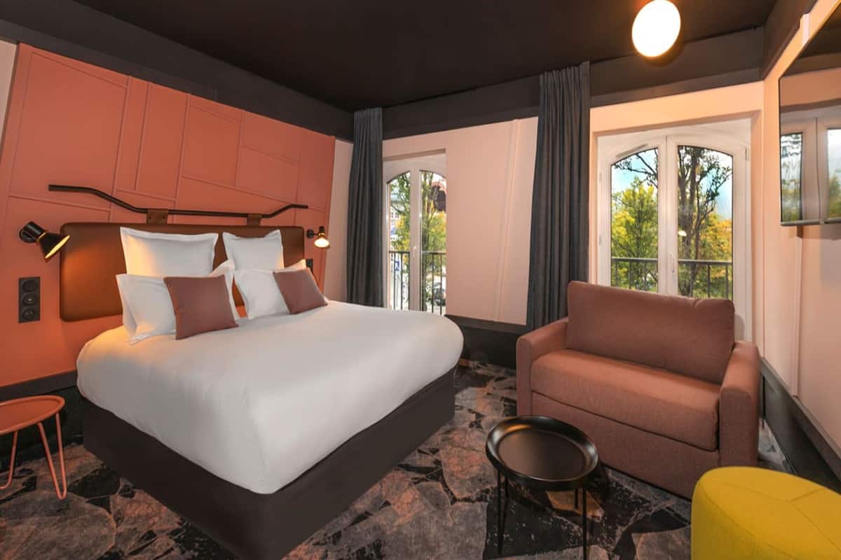A modern hotel room with a large bed, a sofa, a small table, and a view of the outdoors through the windows.