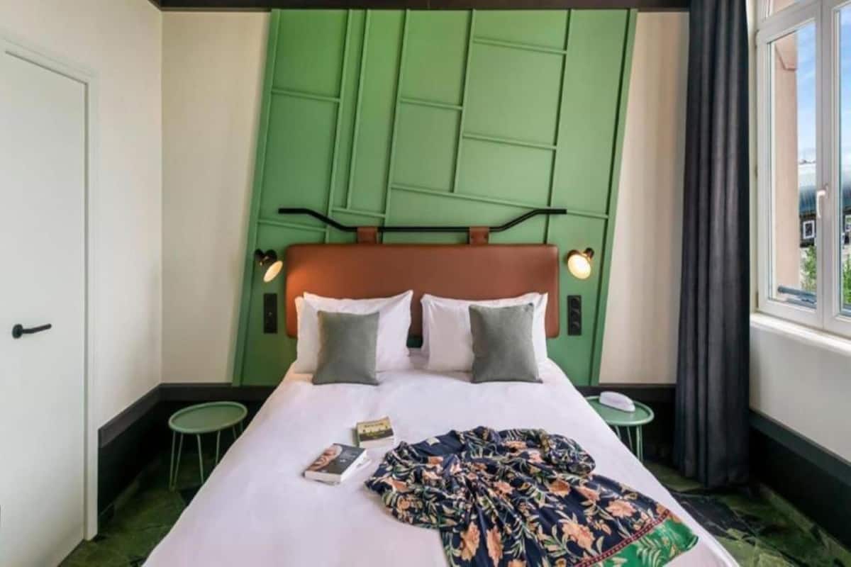 A hotel room with a bed, green headboard, two bedside tables, a window with curtains, and a view of the outside.