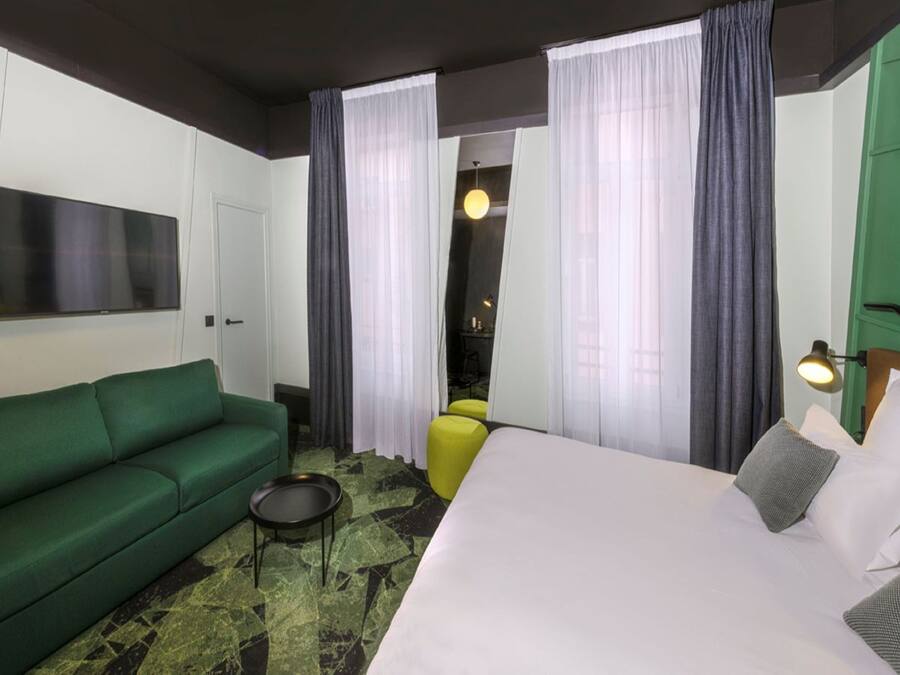 A hotel room with a green sofa, a bed with white bedding, a small round table, and a yellow chair.