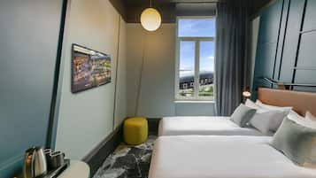 Ein Hotelzimmer mit zwei Betten, einem großen Fenster mit Blick auf die Stadt, einem runden, gelben Hocker und einem gerahmten Bild an der Wand.