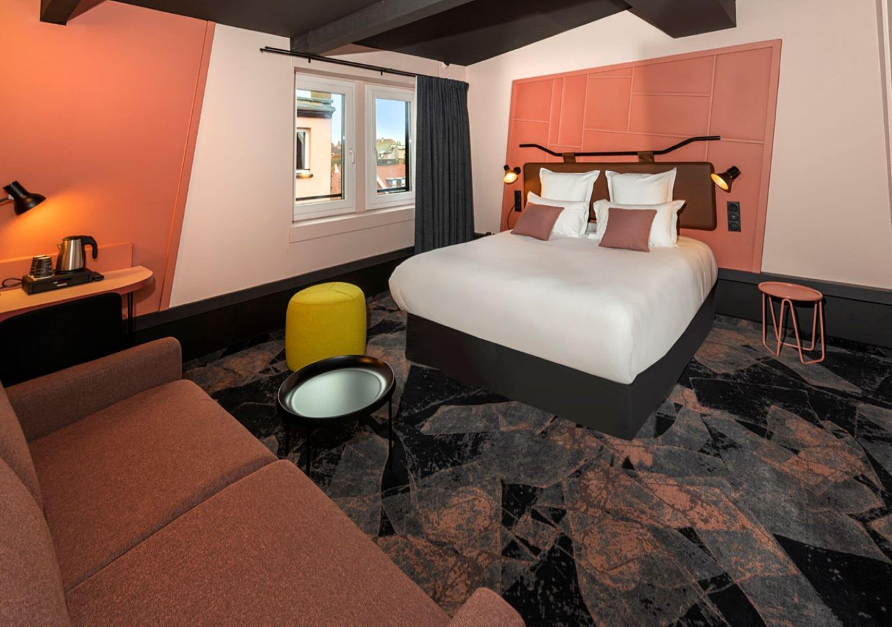 A modern hotel room with a large bed, a sofa, a small table with a kettle, and a view of the city through the window.