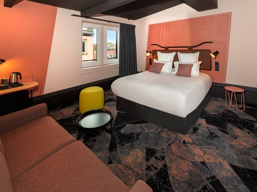 A modern hotel room with a large bed, a sofa, a small table with a kettle, and a view of the city through the window.