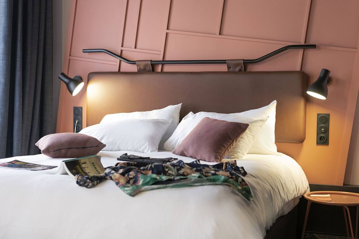 A neatly made bed with pillows, a book, and a blanket. There are two wall-mounted lamps and a small table beside the bed.