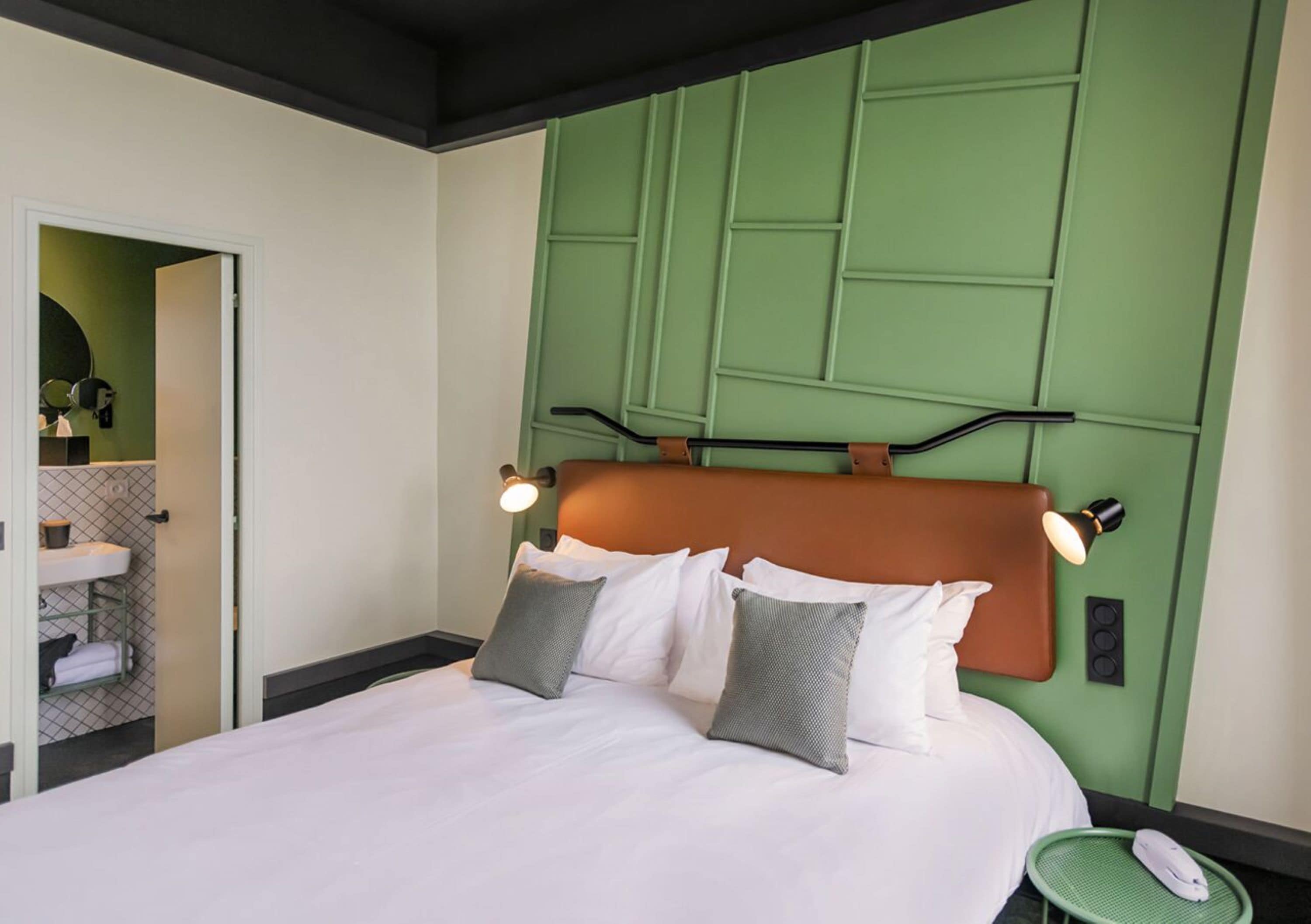 A hotel room with a bed, a green headboard, a bedside table, and a small green chair.