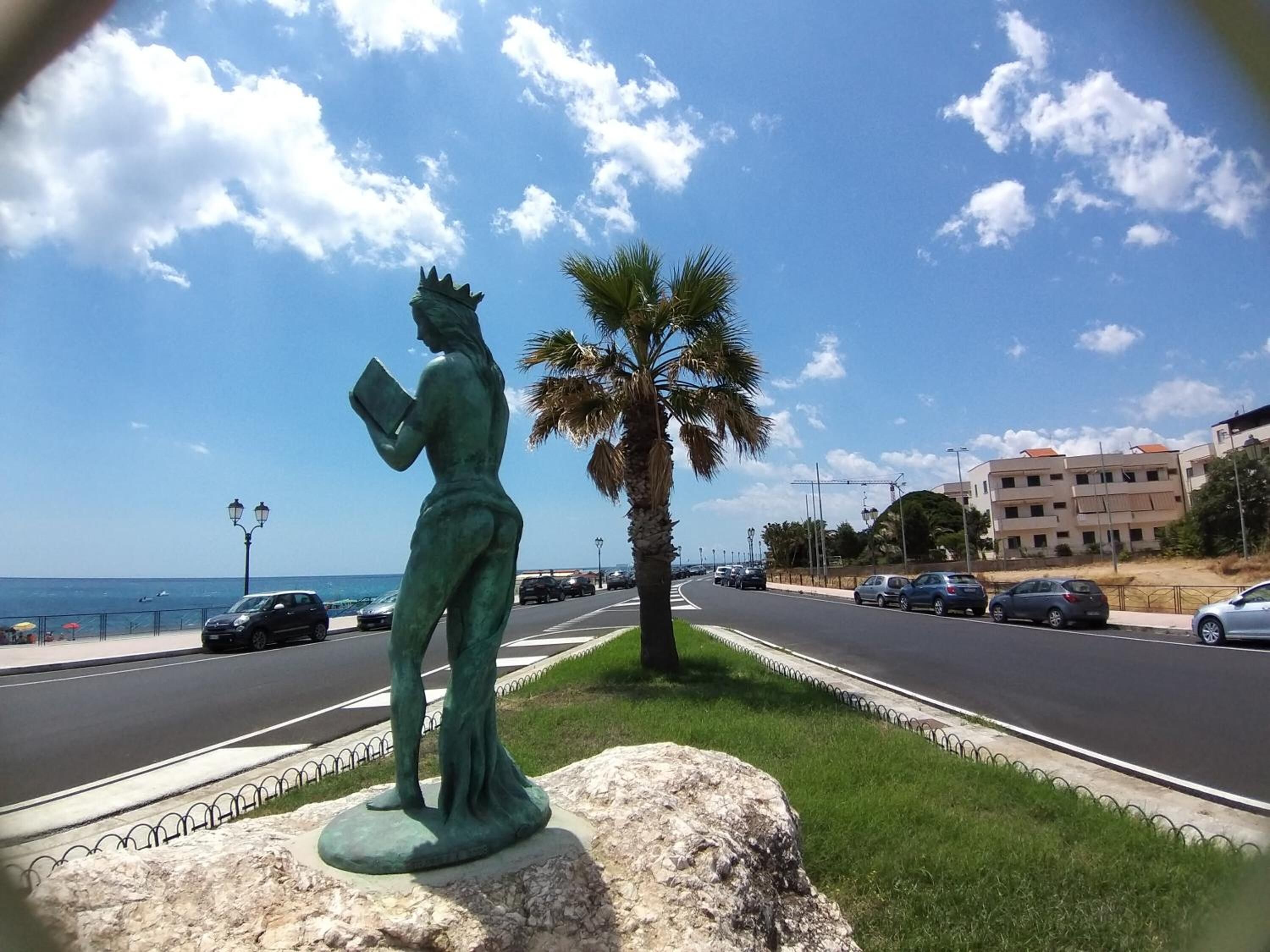 Una estatua sobre un pedestal al borde de la carretera, palmeras, cielo despejado y un camino con coches.