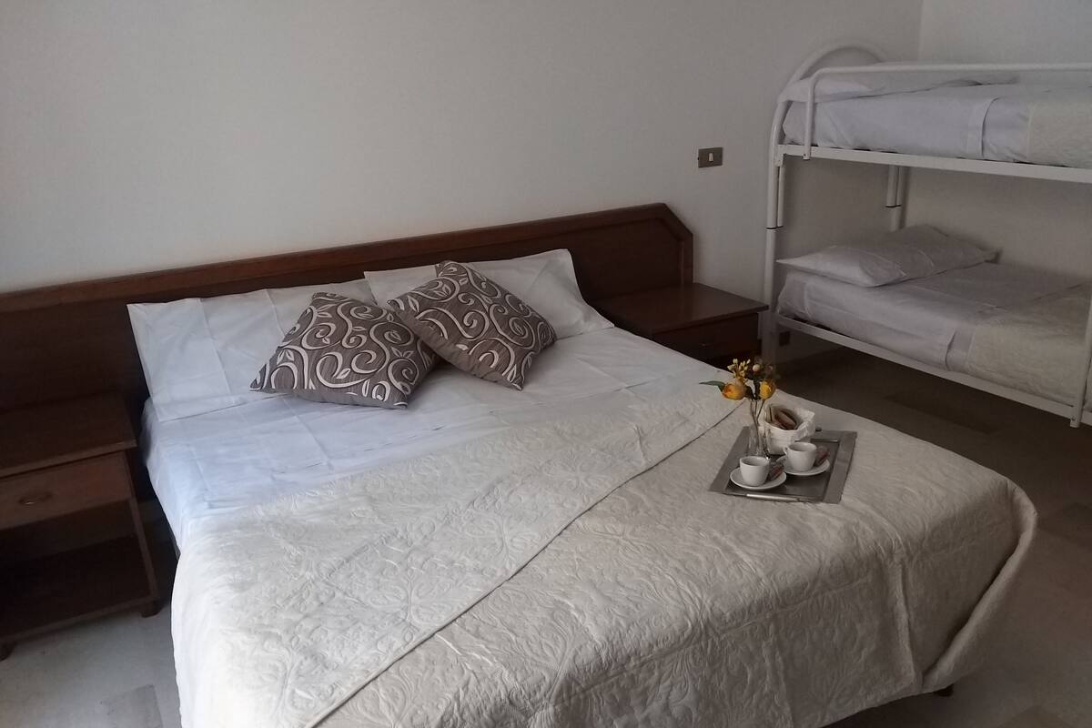 Una cama bien hecha con almohadillas estampadas, una mesita de noche y una cama litera al fondo.