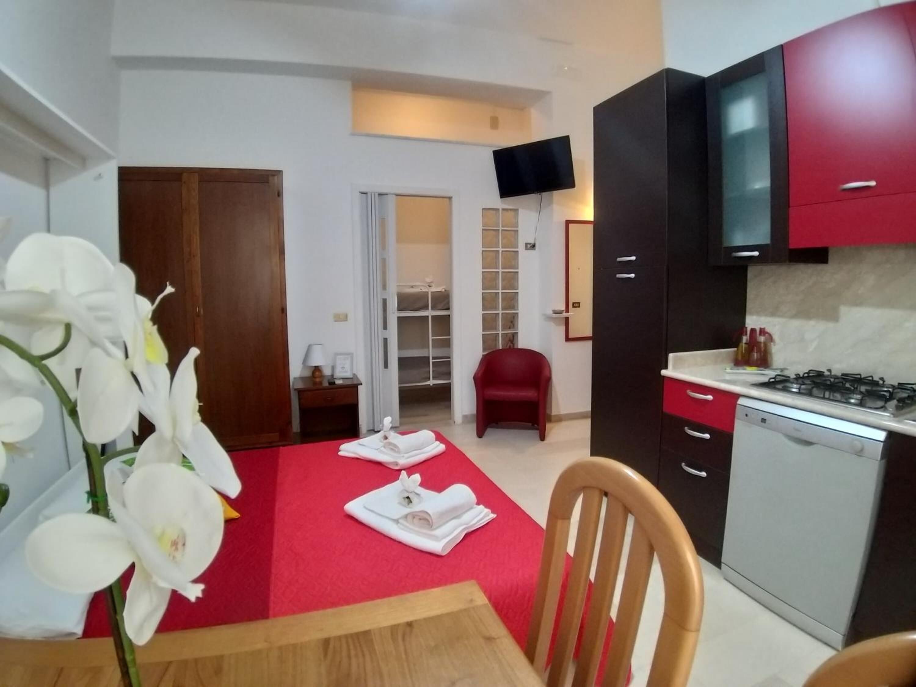 A compact hotel room with a kitchenette, a dining area with a table and chairs, a red carpet, and a bathroom visible through an open door.
