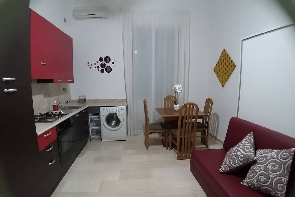 Cocina compacta con lavadora, comedor con mesa y sillas, y sofá rojo con cojines estampados.