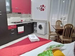 Ett kompakt hotellrum med ett litet kök, en tvättmaskin, en matplats med trästolar och en säng med ett rött täcke.