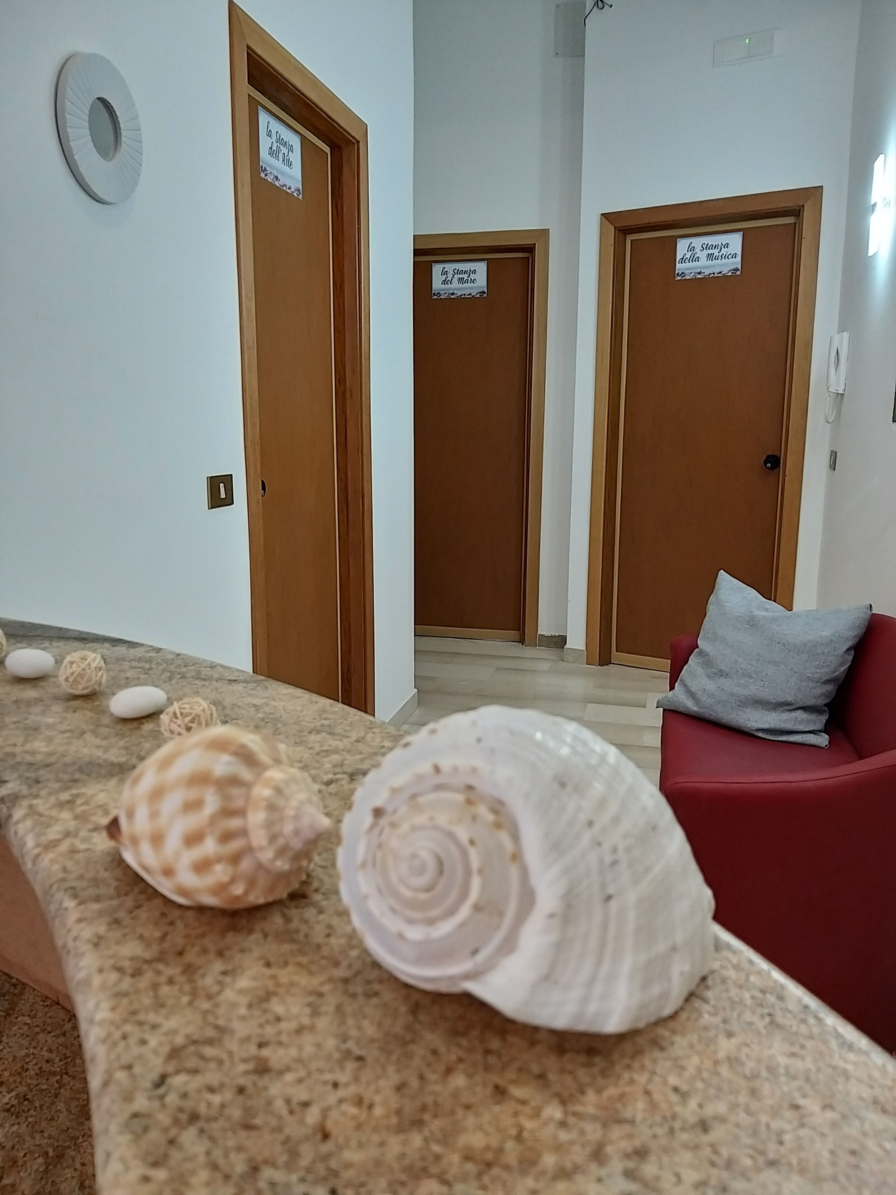 Corredor de hotel com portas de madeira, uma poltrona vermelha e um balcão de mármore com conchas.
