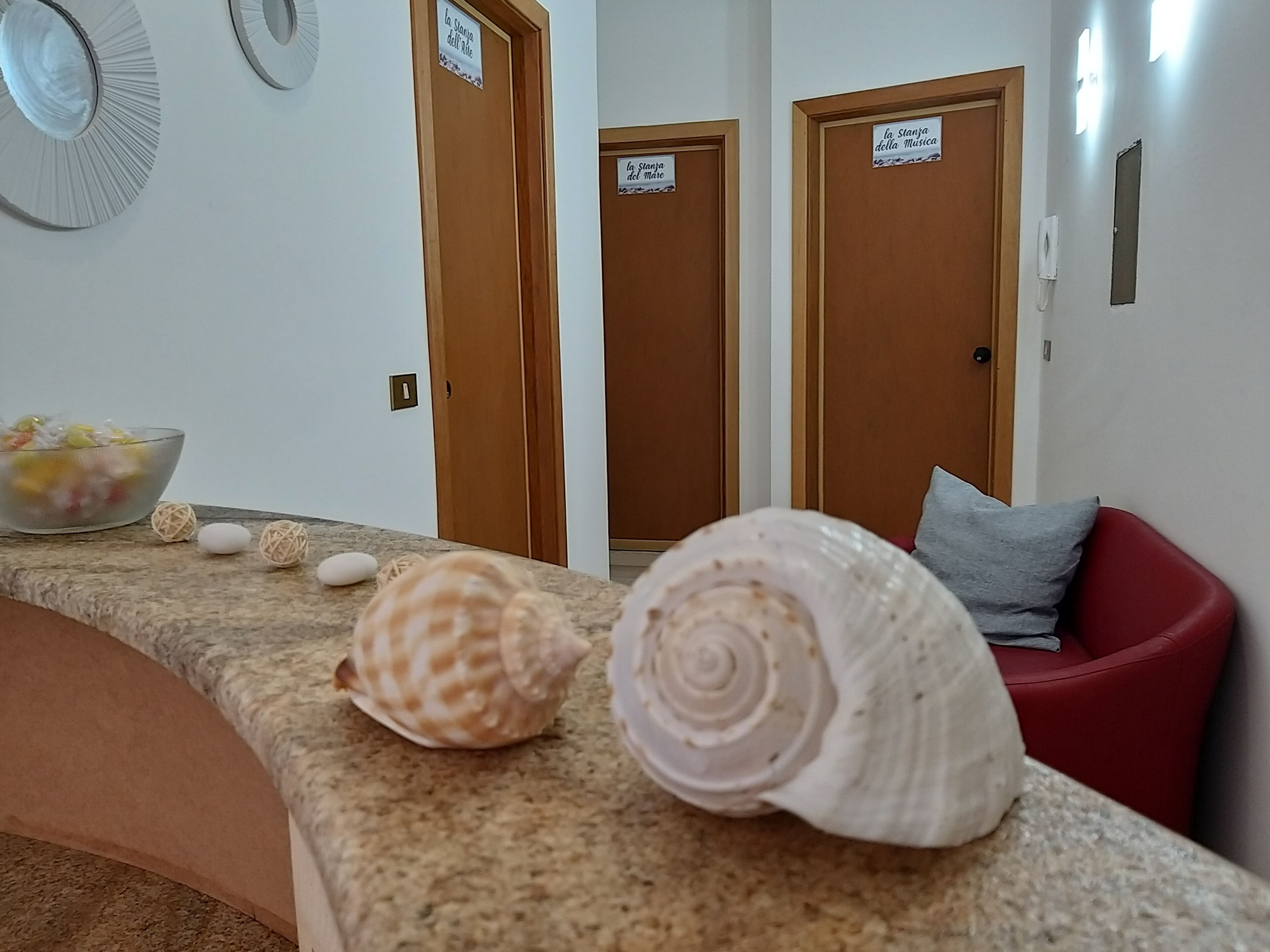 A room with a curved bench, a bowl of fruit, and three doors labeled "la Stanza dell' Aria," "la Stanza del Mare," and "la Stanza della Musica."