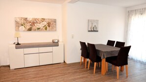 A dining area with a table and chairs, a white cabinet with a lamp, and two wall-mounted artworks. - Ferienwohnungen am Sonnenplatz (Rust)