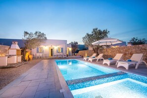 A swimming pool with lounge chairs, a stone wall, and a building with lights.