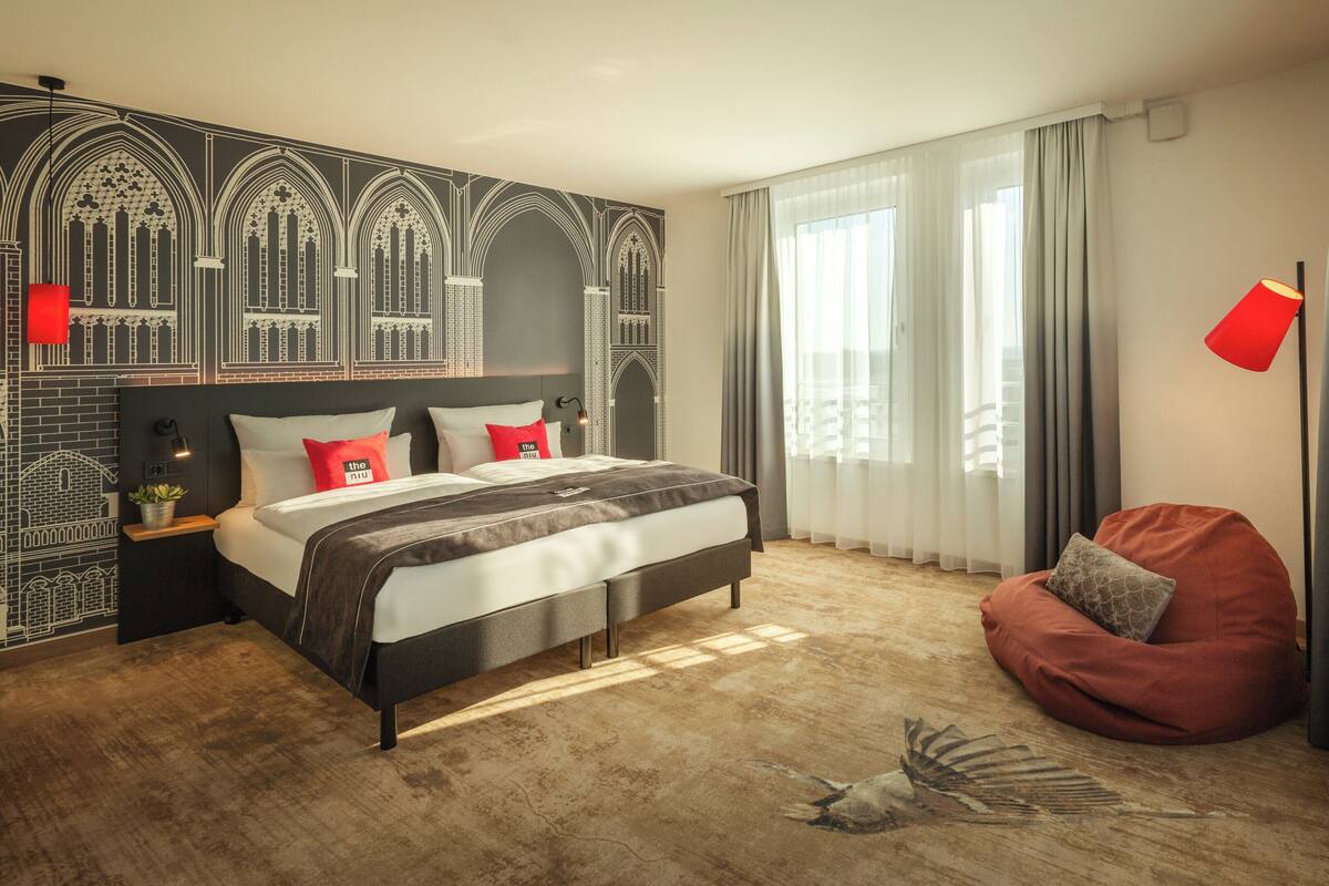 Une chambre d’hôtel moderne avec un grand lit, un fauteuil en forme de sac à haricots rouge et une fresque murale décorative.