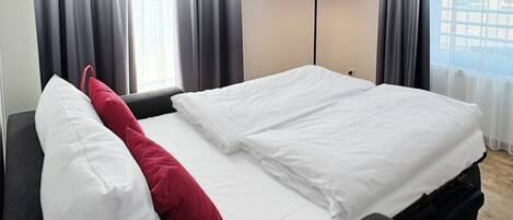 Quarto de hotel com cama, abajur vermelho e vista para a cidade pela janela.
