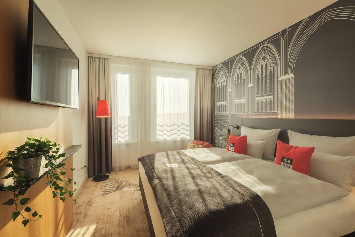 Une chambre d’hôtel avec un grand lit, un téléviseur à écran plat fixé au mur, une plante en pot et une vue sur un immeuble par la fenêtre.