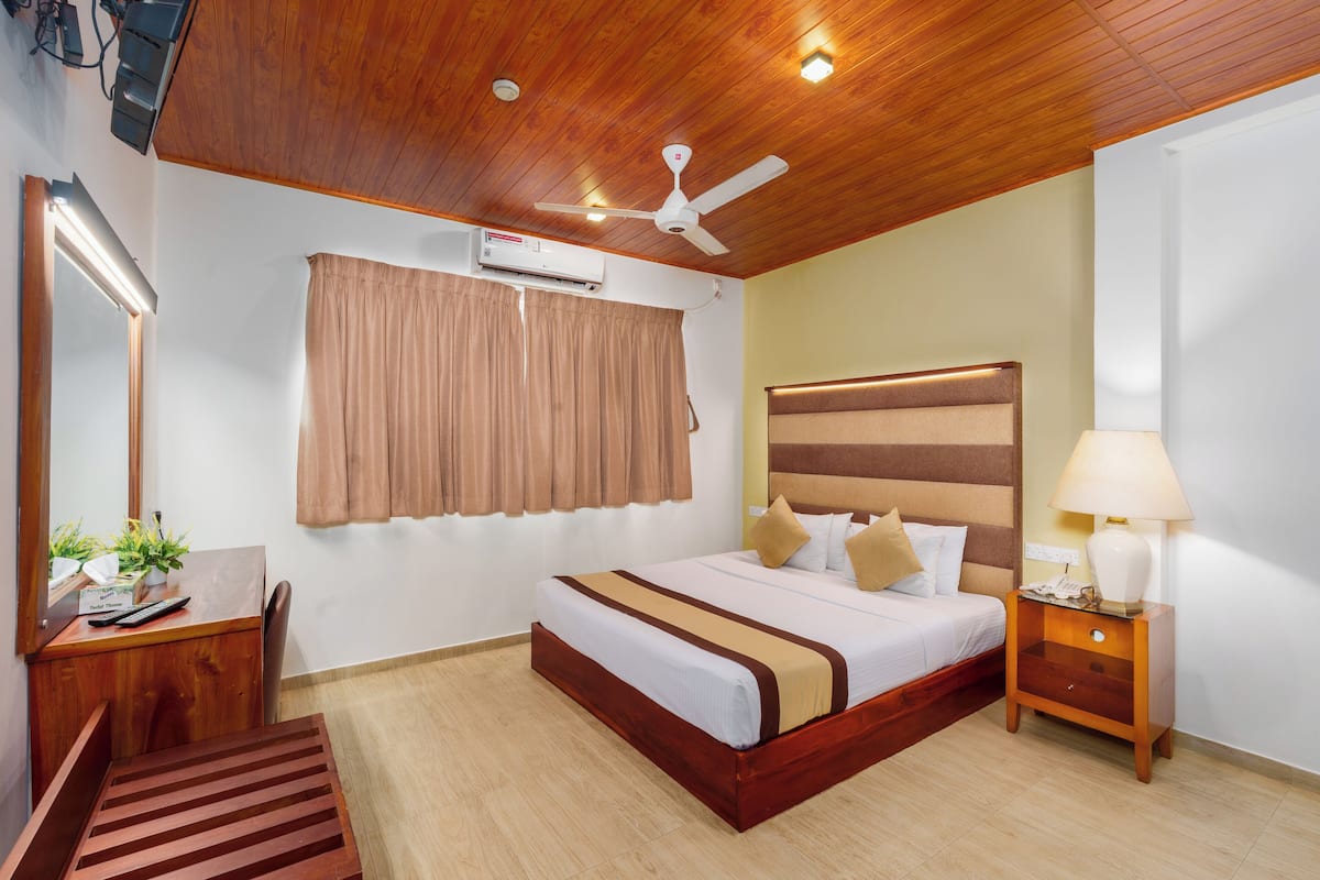 Habitación de hotel con techo de madera, cama con cabecera a rayas, escritorio con espejo y mesita de noche con lámpara.