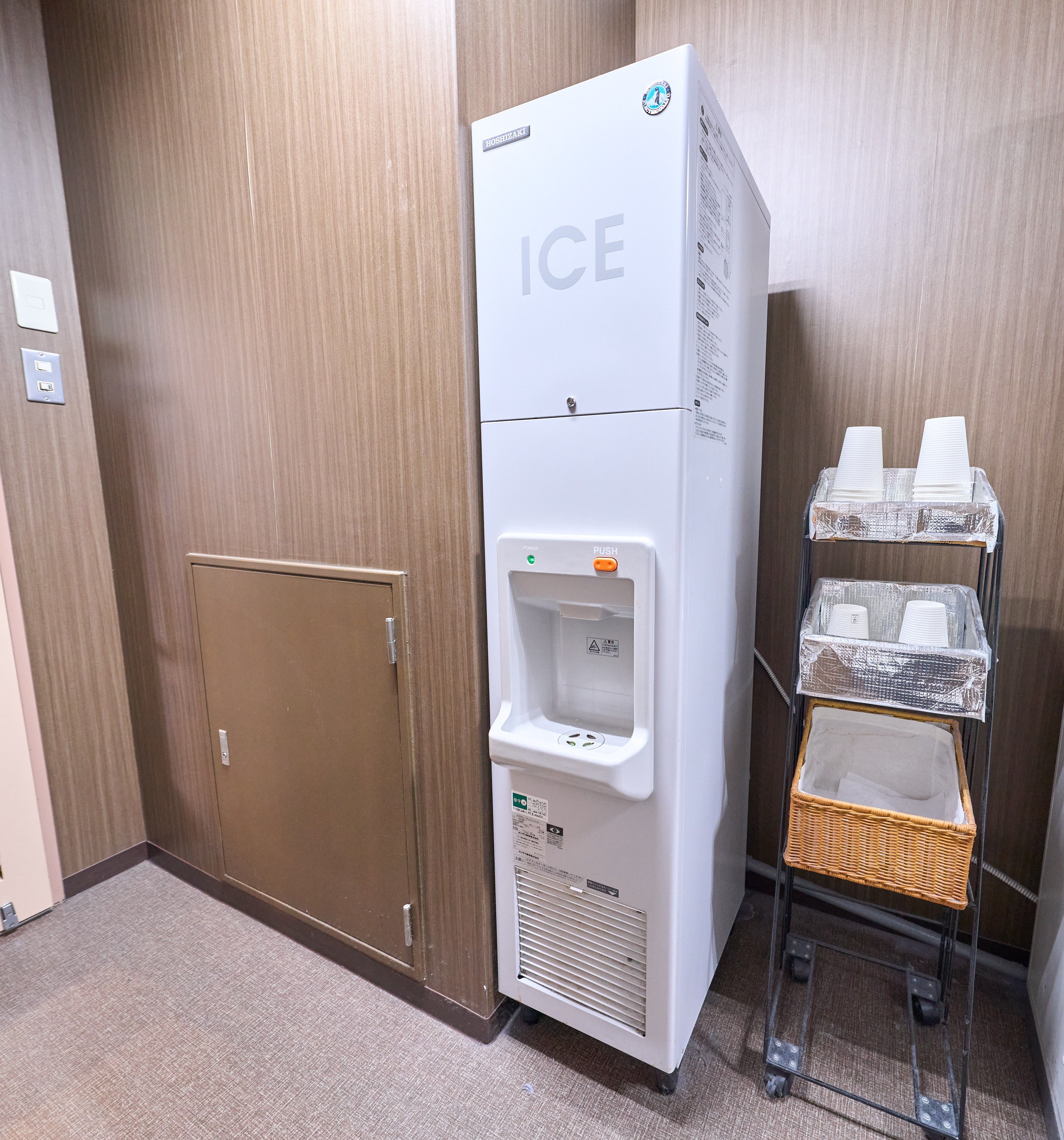A hotel room with a white ice dispenser, a brown door, and a small wicker basket on a cart.
