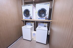 A laundry facility with two front-loading washing machines and two dryers, all with control panels and safety labels. - Smile Hotel Sasebo (Sasebo)