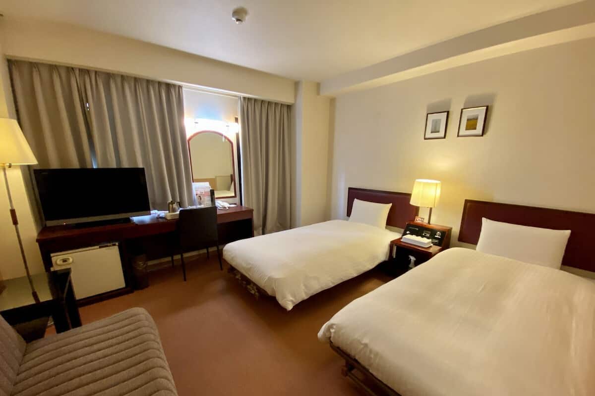 A hotel room with two beds, a desk with a TV, a chair, and a small table with a lamp.