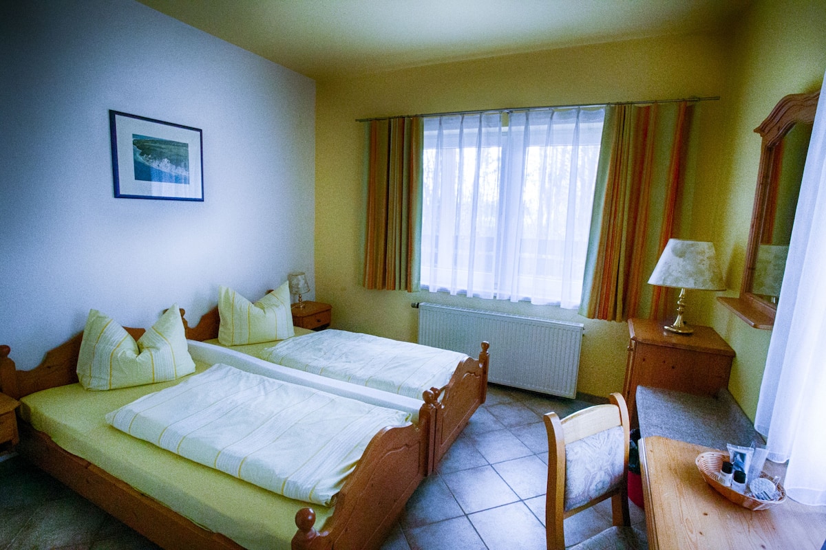 A double bed with a wooden frame, a bedside table with a lamp, and a window with curtains.