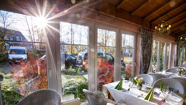 A dining area with wicker chairs and tables set for a meal, large windows with a view of parked cars and trees, and a wooden ceiling. - Hotel Rosengarten (Naunhof)