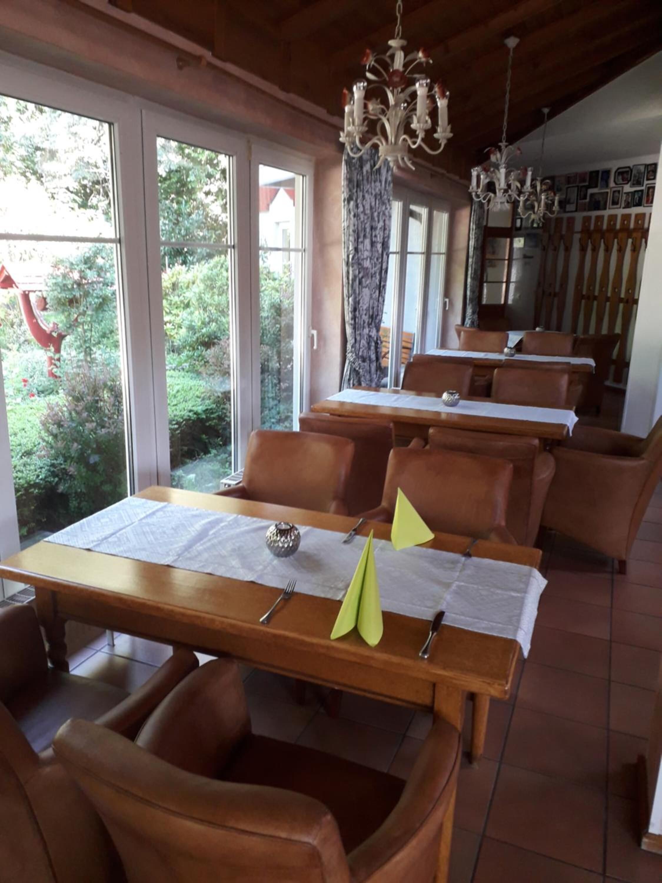 A dining area with wooden tables and chairs, large windows with curtains, and chandeliers.