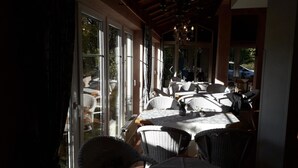A sunlit patio with wicker furniture and a table, large windows, and a chandelier. - Hotel Rosengarten (Naunhof)