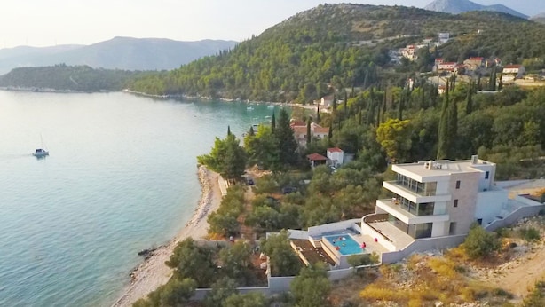 A modern multi-story building with a rooftop pool, situated on a hillside overlooking a calm sea with a boat in the distance.