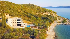 A modern two-story building with a pool, surrounded by lush greenery and a coastal view.