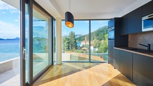 A modern kitchen with a large glass door leading to a balcony overlooking a scenic view of the sea and hills.