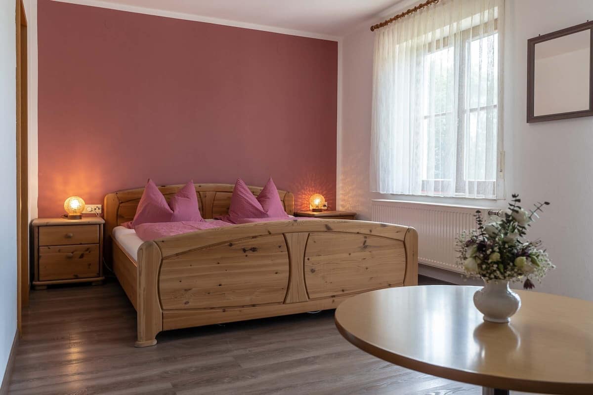 A bedroom with a wooden bed, two bedside tables with lamps, a round table with a vase, and a window with curtains.