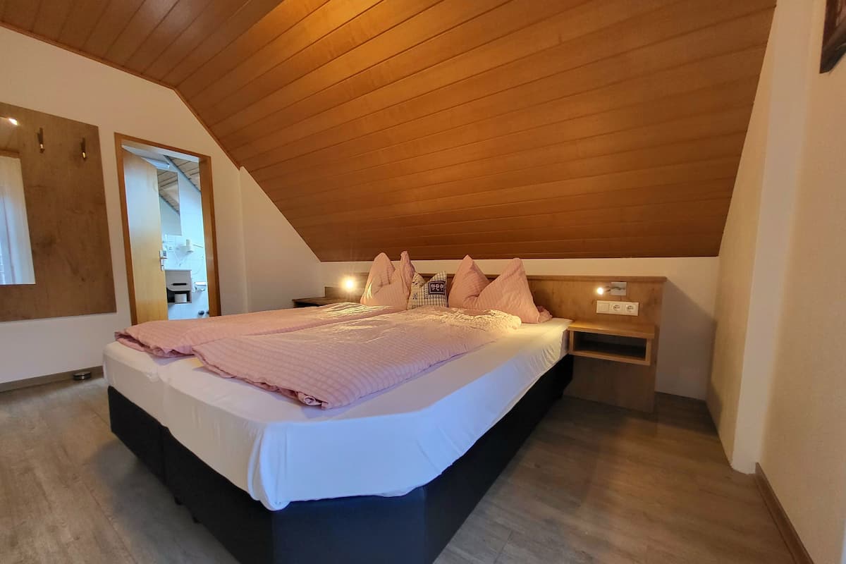 A bedroom with a sloped ceiling, a bed with a pink bedspread, a bedside table, and a nightstand.