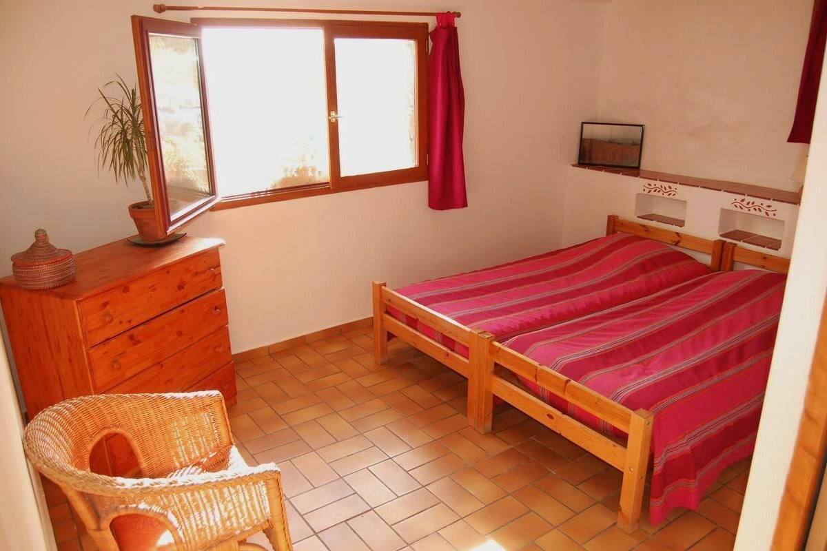 A bedroom with a wooden bed, a dresser, a wicker chair, a window with curtains, and a small shelf with a picture.