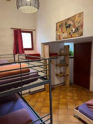 Une chambre de dortoir avec des lits superposés, une fenêtre avec des rideaux rouges, une étagère fixée au mur et une porte.
