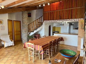 Un espace repas avec une longue table, des chaises, un banc en bois, un pilier en pierre, un escalier et, au fond, un coin cuisine.