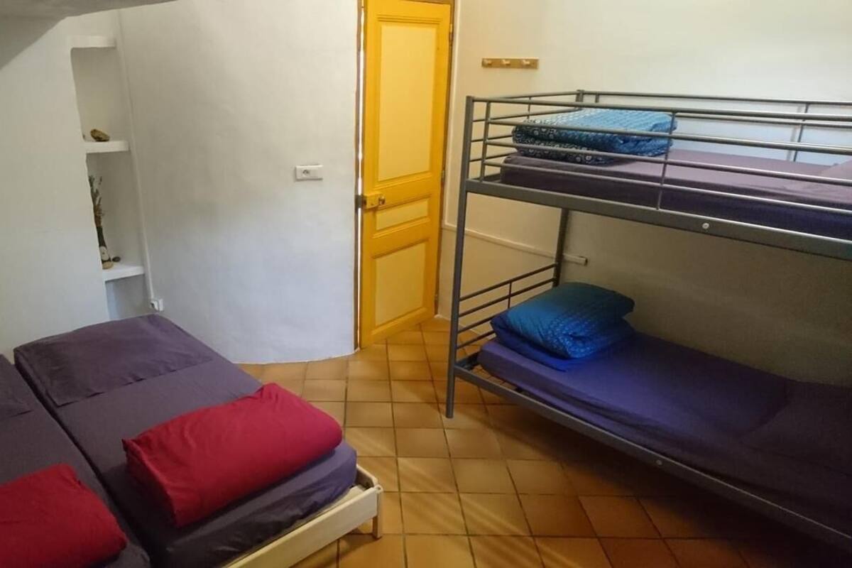 A dormitory room with two beds, one with red and blue bedding and the other with blue bedding. There is a bunk bed with blue bedding on the right. A door is visible in the background, leading to another room.