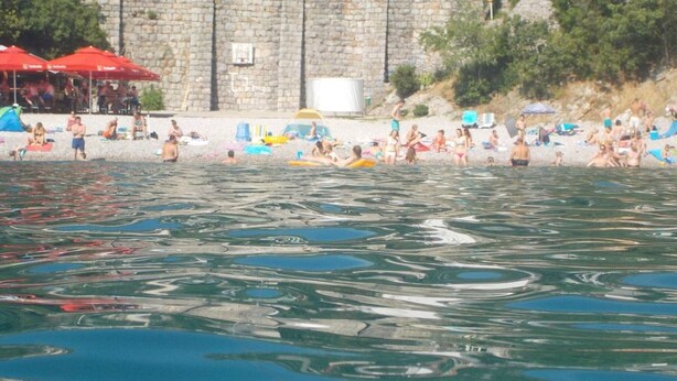 Ein Strand mit badenden und sonnenden Personen, einer Steinmauer und roten Sonnenschirmen.