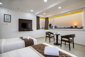 Habitación de hotel con dos camas, un televisor, una mesita con una silla y vistas al atardecer a través de un ventanal grande.