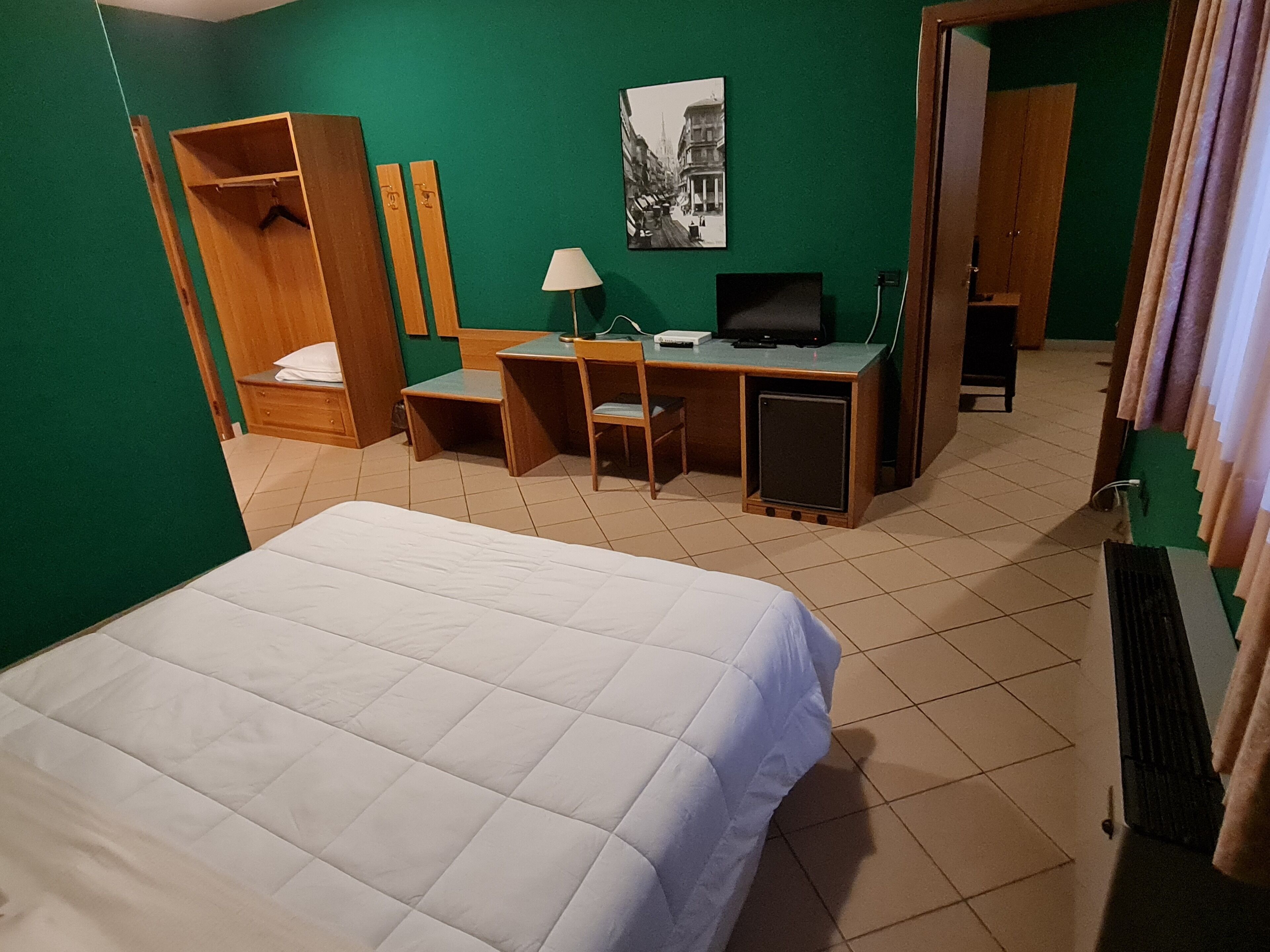 Habitación de hotel con cama, escritorio con computadora, silla, armario y un cuadro en la pared.